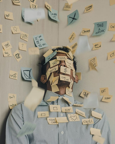 Homme en chemise bleu adossé à un mur, le visage recouvert de post-it l'incitant à prendre du repos